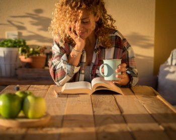 women at table with hand on her chin looking down at open book with coffee in the other hand